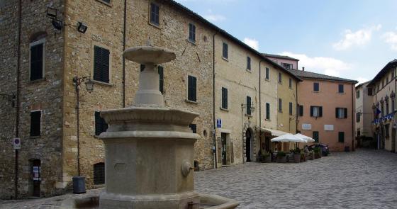 Piazza tranquilla con fontana storica, caffè all'aperto e case in pietra.